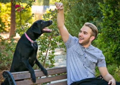 Outdoor senior portrait session of a boy and his black lab on a bench with greenery all around them while he holds a treat up for his dog