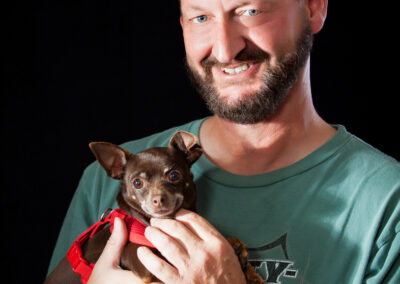 Man holding his brown chihuahua to his chest against a black backdrop