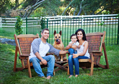 Backyard family portrait with husband and wife sitting next to each other on wooden chairs with their baby in their lap and their dog posed in between them