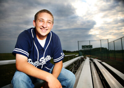 Senior boy smiling sitting on the bleachers of a baseball field with the view of the sky behind him