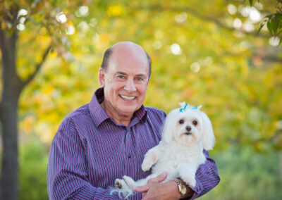 Older man holding his little white dog with blue bows in its hair in our portrait park in Citrus Heights, CA