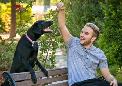 Young man sitting on a wooden bench outside holding up a treat for his black Labrador dog