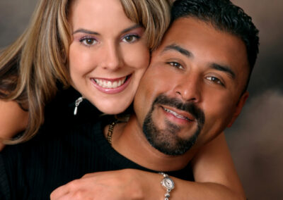 Woman posed lovingly embracing man from behind with both smiling at the camera and a brown backdrop as the background