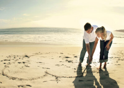 Couple in matching white shirt and denim outfits drawing a heart together in the sand at Carmel beach, CA