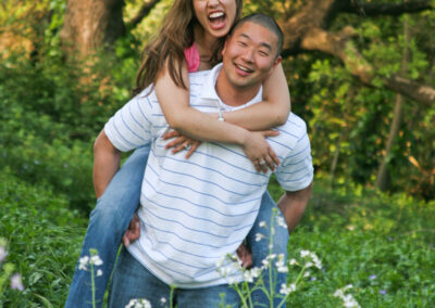 Couple posed piggyback with the woods in the background with lots of greenery and small white flowers surrounding them while smiling at the camera