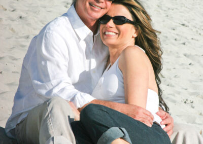 Couple holding each other smiling at camera while sitting on a sandy beach wearing matching outfits