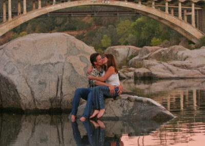 Couple lovingly posed sitting atop a large rock in Lake Natoma in Folsom, CA with the bridge behind them