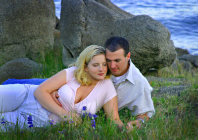 Couple lying down embracing each other in gorgeous greenery and purple flowers with large rocks and the ocean calmly behind them.