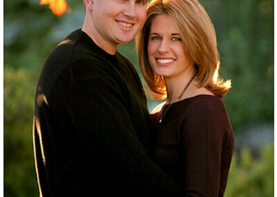 Couple smiling at the camera while holding each other in a loving embrace outside under the fall trees